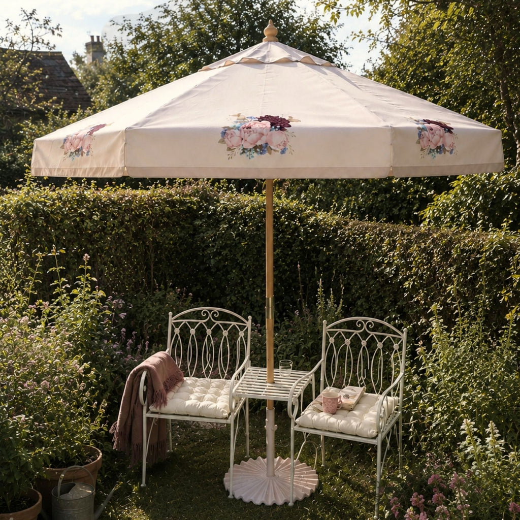 A cream-colored outdoor patio umbrella with delicate pink and maroon floral motifs, set in a lush green garden next to two white wrought-iron garden chairs with cream cushions and a small white table.