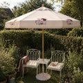A cream-colored outdoor patio umbrella with delicate pink and maroon floral motifs, set in a lush green garden next to two white wrought-iron garden chairs with cream cushions and a small white table.
