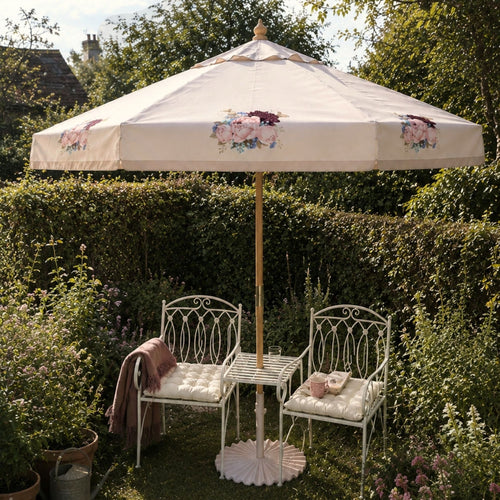 A cream-colored outdoor patio umbrella with delicate pink and maroon floral motifs, set in a lush green garden next to two white wrought-iron garden chairs with cream cushions and a small white table.