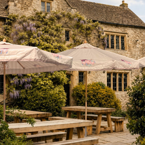 Outdoor patio seating with large cream patio umbrellas and wooden picnic tables set against a stone building covered in green ivy and purple wisteria flowers.