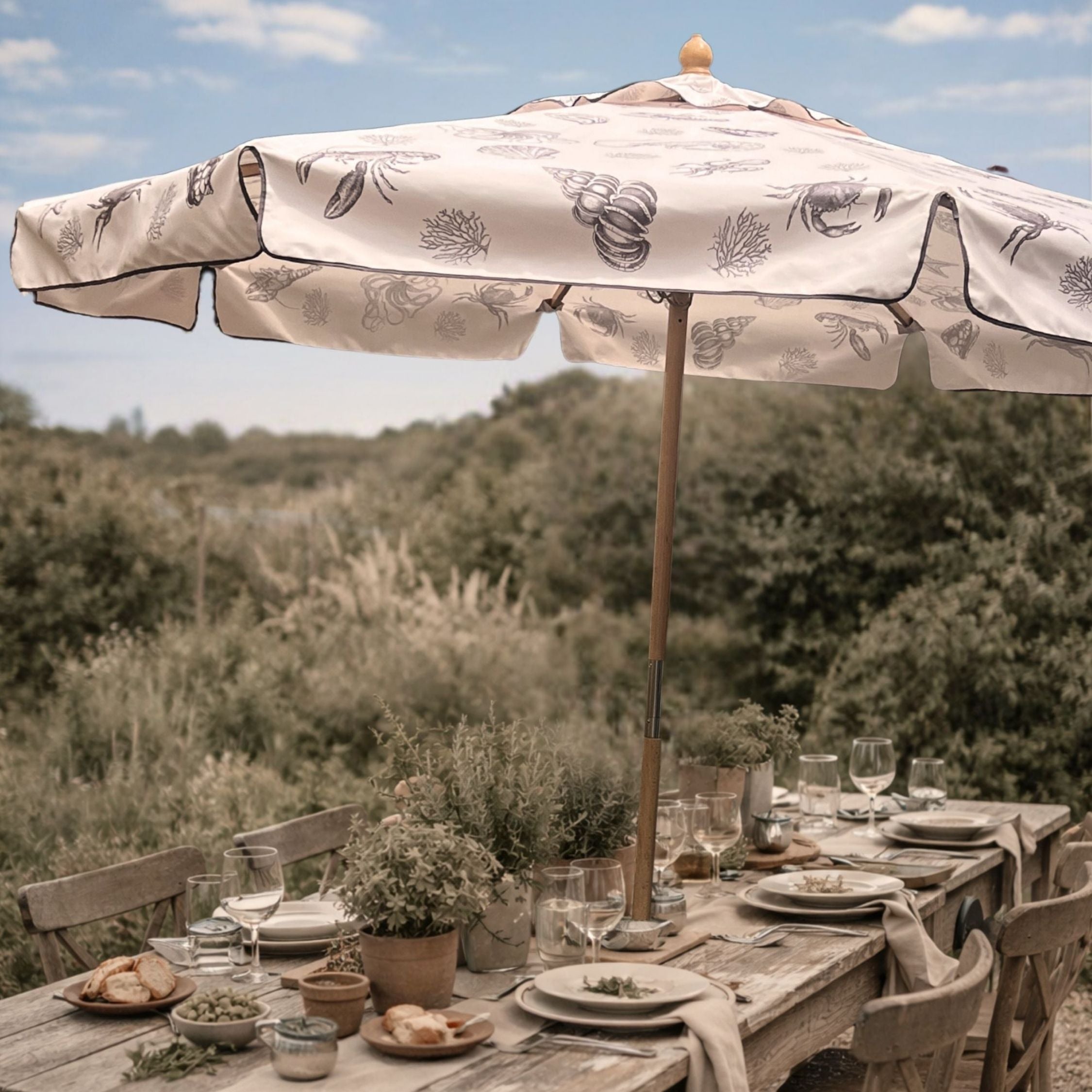 A large patio umbrella with a cream fabric canopy and dark botanical line art print stands over an outdoor dining table set with plates, glasses, and plants, in a natural garden setting.