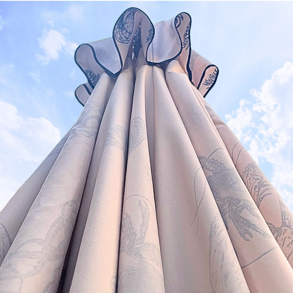 A close-up, low-angle view of a cream canvas parasol canopy with navy piping around the air vent section,  set against a blue sky with clouds.