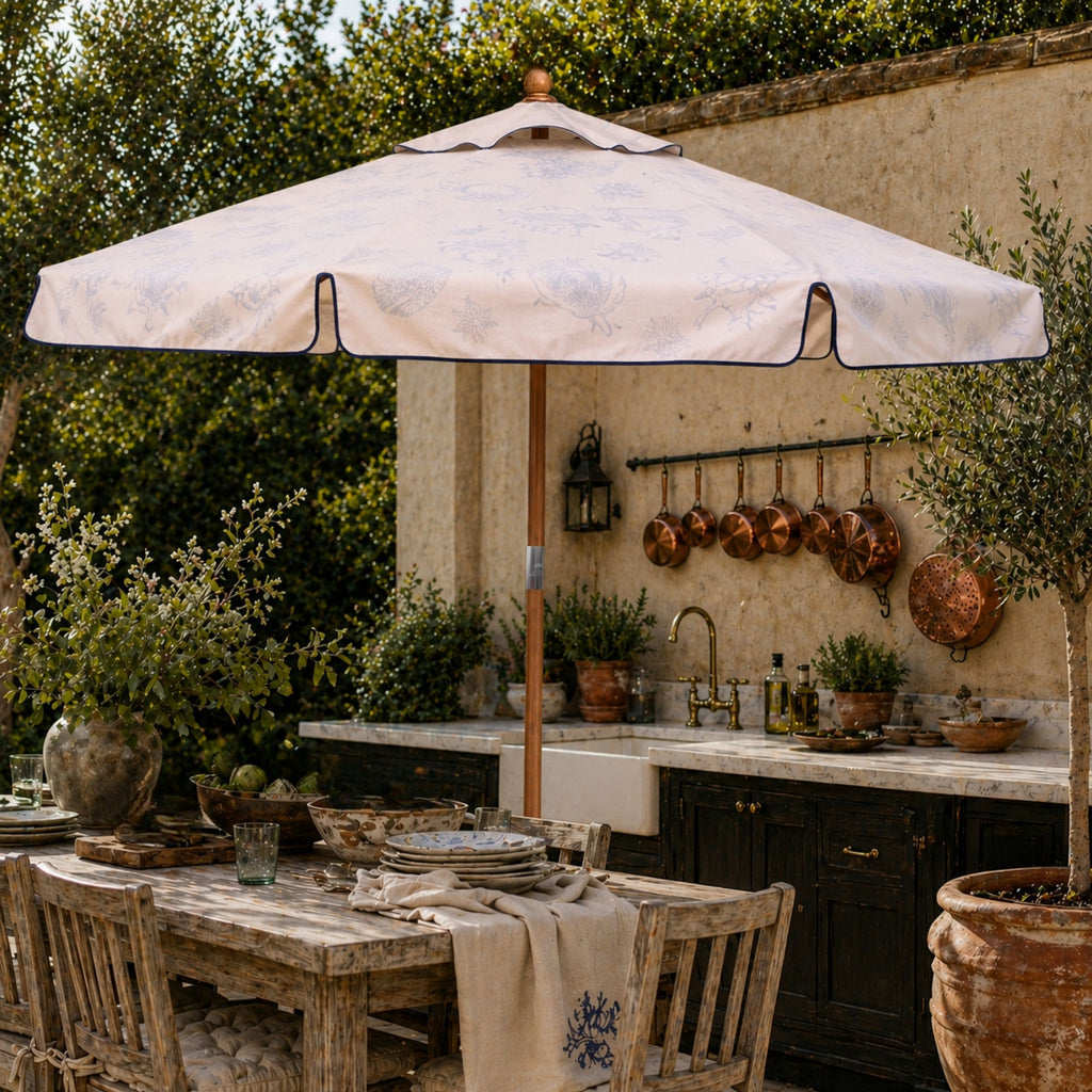 A large outdoor patio umbrella with a light beige outer canopy with navy trim. The inside of the canopy is designed with maritime illustrations. The parasol is shading a rustic wooden dining table in a styled outdoor kitchen setting.