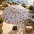 A large patio umbrella with a white canopy and blue nautical sea creature print, set on a stone terrace overlooking the ocean with outdoor seating nearby.