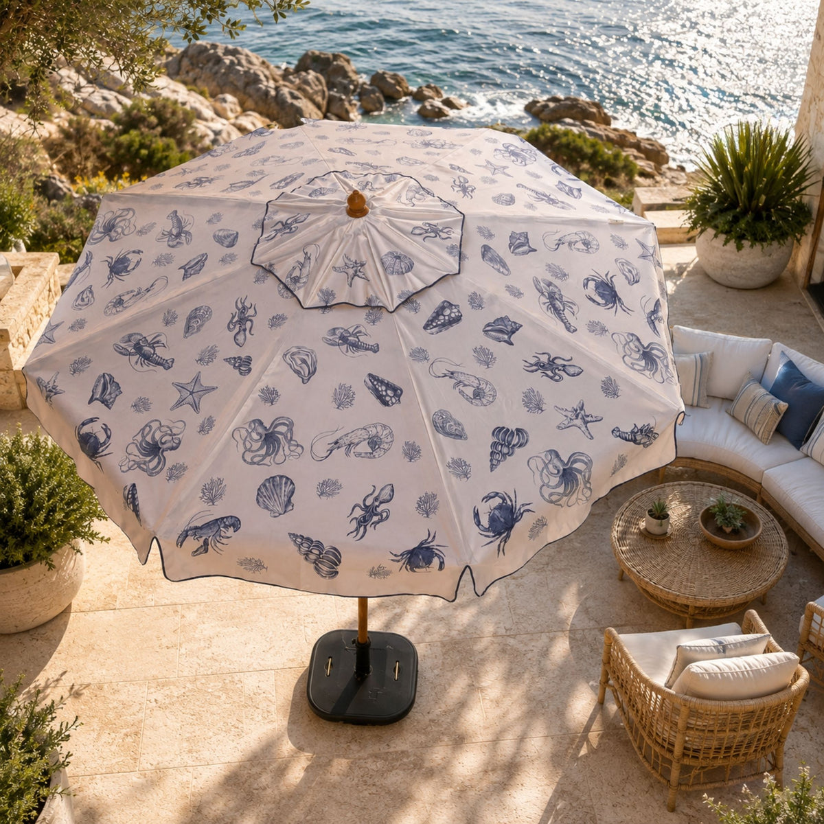 A large patio umbrella with a white canopy and blue nautical sea creature print, set on a stone terrace overlooking the ocean with outdoor seating nearby.