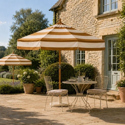 A large striped patio umbrella in  caramel and cream stripe stands over a small round metal table and two matching chairs on a stone patio in front of a rustic stone house.