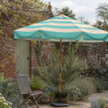 A large patio umbrella with teal and cream stripes stands in a garden courtyard beside a wooden chair, with a brick wall and potted plants in the background.