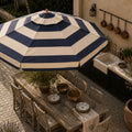 A large navy blue and cream striped patio umbrella shades a wooden dining table set with chairs, plates, and bowls on a stone patio next to an outdoor kitchen sink.