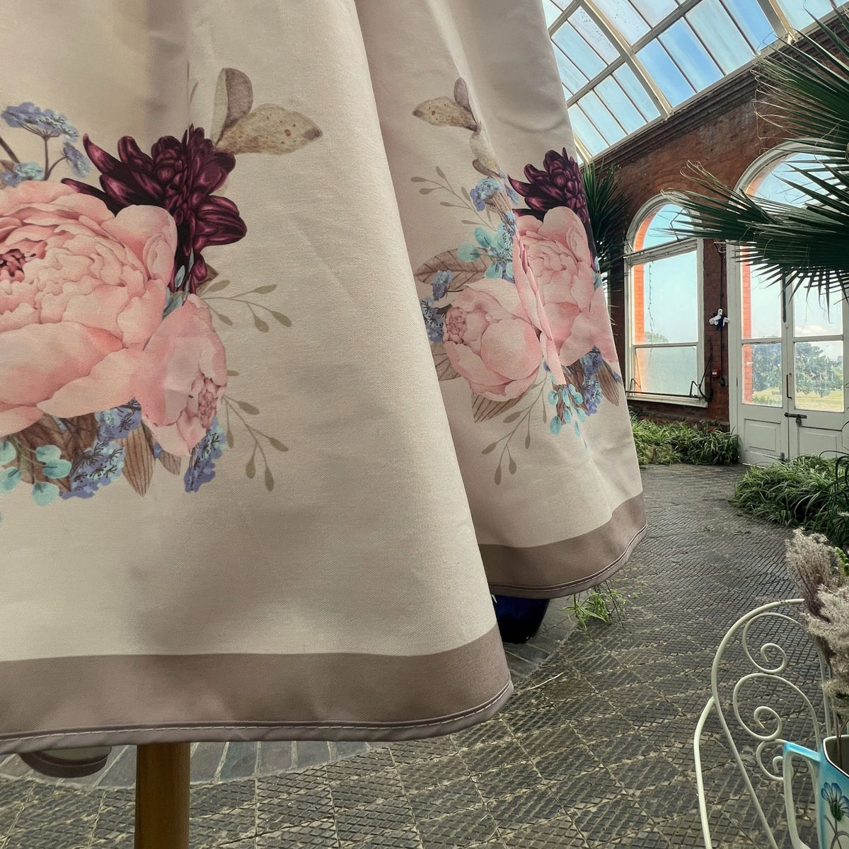 A close-up of a cream-coloured solid wood garden parasol,  featuring a large pink peony and dark burgundy floral print, with a taupe border, standing in a orangery interior.