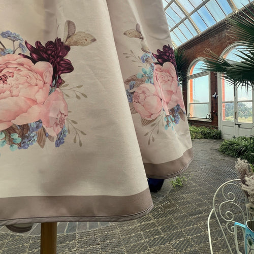 A close-up of a cream-coloured solid wood garden parasol,  featuring a large pink peony and dark burgundy floral print, with a taupe border, standing in a orangery interior.